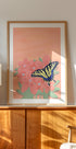 een ingelijst schilderij van een vlinder op een roze bloem, tentoongesteld op een houten kast of plank.