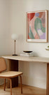 een minimalistische werkruimte met een houten bureau, een geweven stoel, een lamp en een ingelijst abstract schilderij aan de muur.