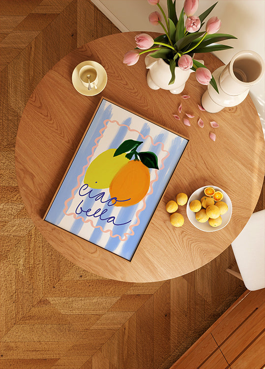 een houten tafel met een ingelijst kunstwerk van twee citroenen, een schaal met fruit en een vaas met roze bloemen.