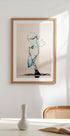 een ingelijst kunstwerk van een waterdruppel die in de lucht hangt, met een blauw en wit kleurenschema. Het kunstwerk hangt op een witte muur boven een tafel met een witte vaas en een boek.