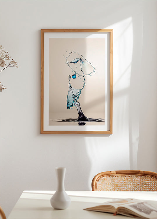 een ingelijst kunstwerk van een waterdruppel die in de lucht hangt, met een blauw en wit kleurenschema. Het kunstwerk hangt op een witte muur boven een tafel met een witte vaas en een boek.