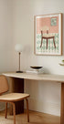 een gezellig hoekje van een kamer met een houten bureau, een geweven stoel, een lamp en een ingelijst kunstwerk van een stoel op een vloer met een patroon.