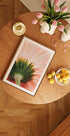 een houten tafel met een ingelijste afbeelding van een bloem, een schaal met fruit en een vaas met roze bloemen.