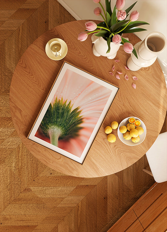 een houten tafel met een ingelijste afbeelding van een bloem, een schaal met fruit en een vaas met roze bloemen.
