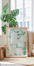 een grote ingelijste print van witte bloemen met groene bladeren, geplaatst op een houten vloer in een lichte, luchtige kamer met grote ramen.