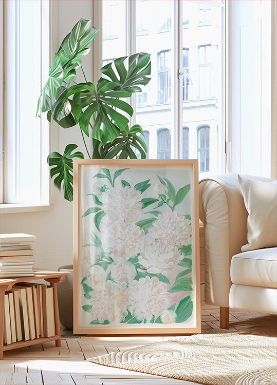 een grote ingelijste print van witte bloemen met groene bladeren, geplaatst op een houten vloer in een lichte, luchtige kamer met grote ramen.