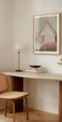een moderne, minimalistische werkruimte met een houten bureau, een geweven stoel, een lamp en een ingelijst schilderij van een huis.