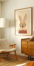 een gezellige woonkamer met een beige fauteuil, een houten ladekast en een ingelijste foto van een konijn met een bril.