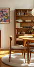 een gezellige en overzichtelijke woonkamer met een houten boekenplank, een ronde houten tafel en een houten stoel. De kamer heeft een warme en uitnodigende sfeer, met een schilderij aan de muur en een schaal met fruit op tafel.